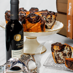 A chocolate babka cake inside a leather book-shaped box, accompanied by a bottle of Cabernet Sauvignon wine and a mask with intricate designs.