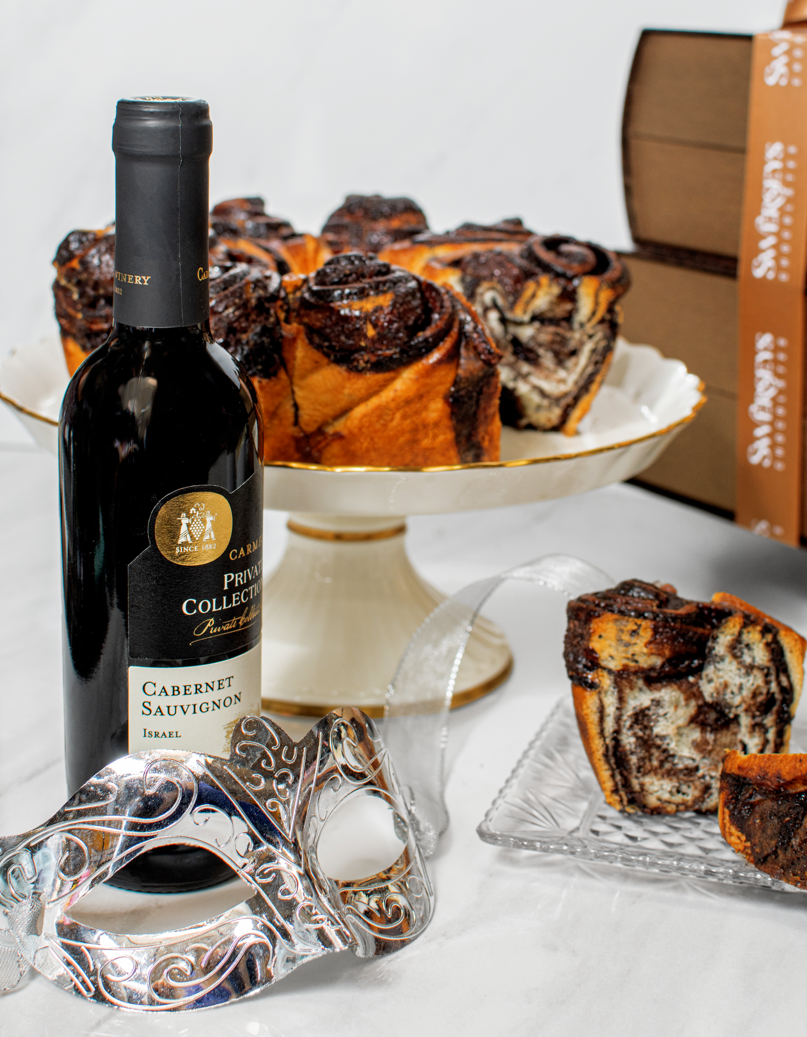 A chocolate babka cake inside a leather book-shaped box, accompanied by a bottle of Cabernet Sauvignon wine and a mask with intricate designs.