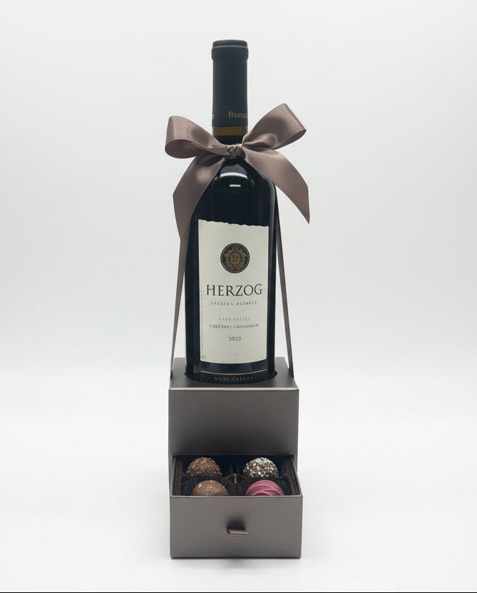 Red wine bottle with a bow and a small box of chocolates on a white surface.