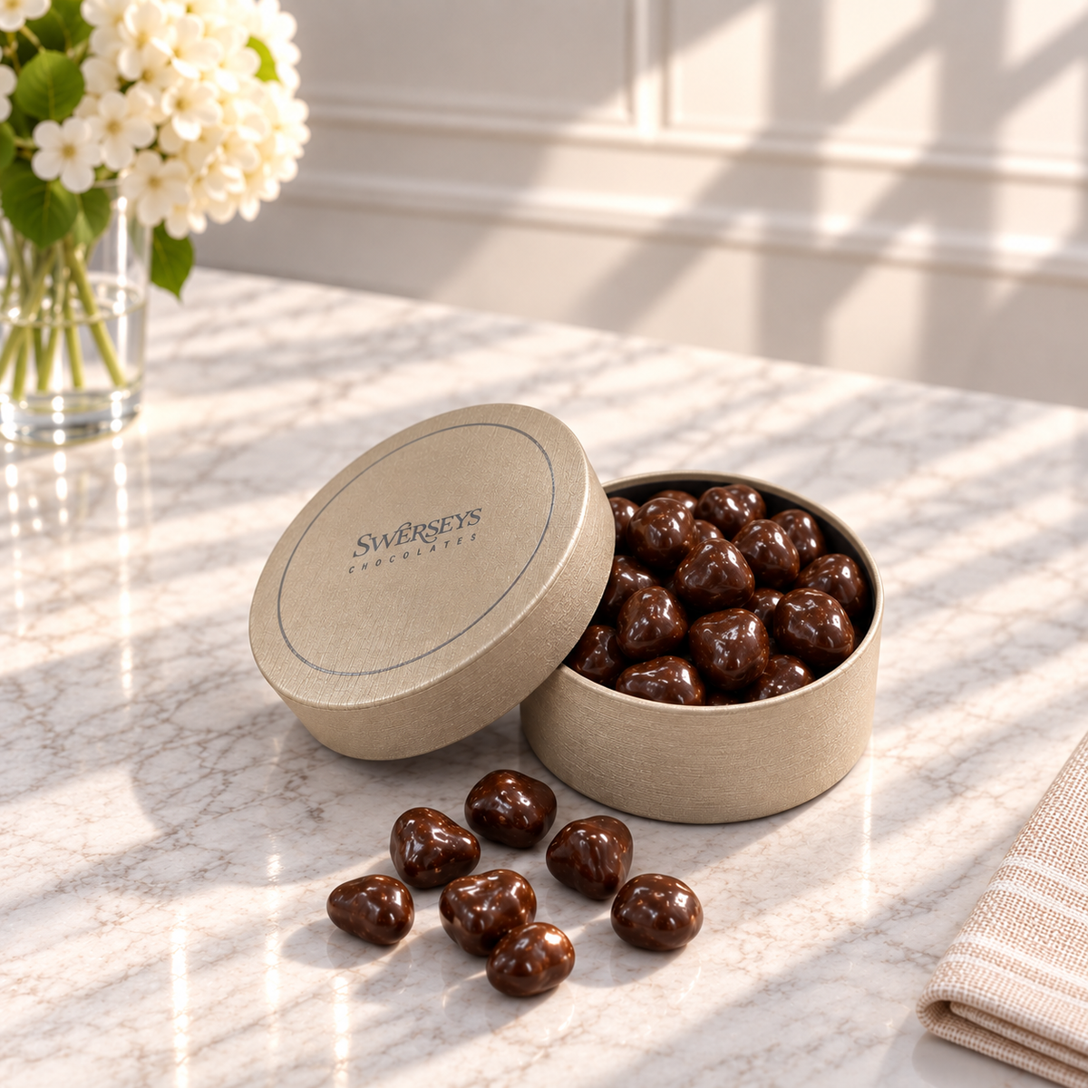Kosherline chocolate espresso beans in a round gift box with lid on a marble table next to white flowers