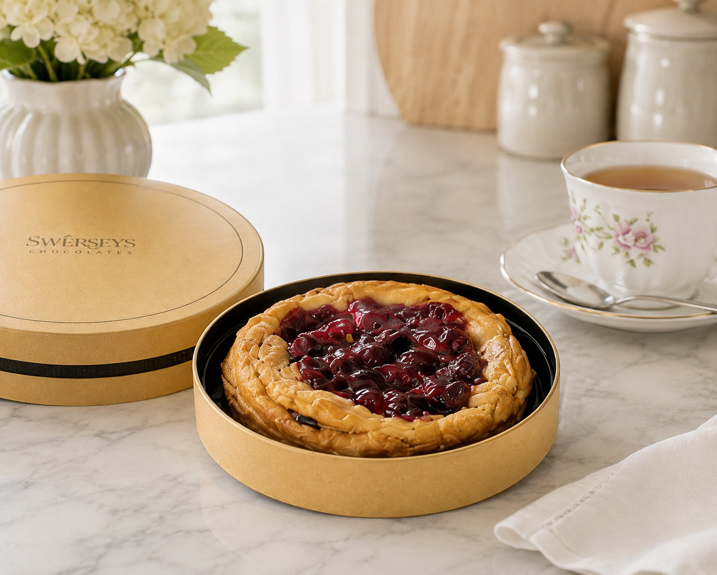 Kosherline Swerseys gourmet fruit mini pie in a round gold gift box on a marble table with a floral teacup.