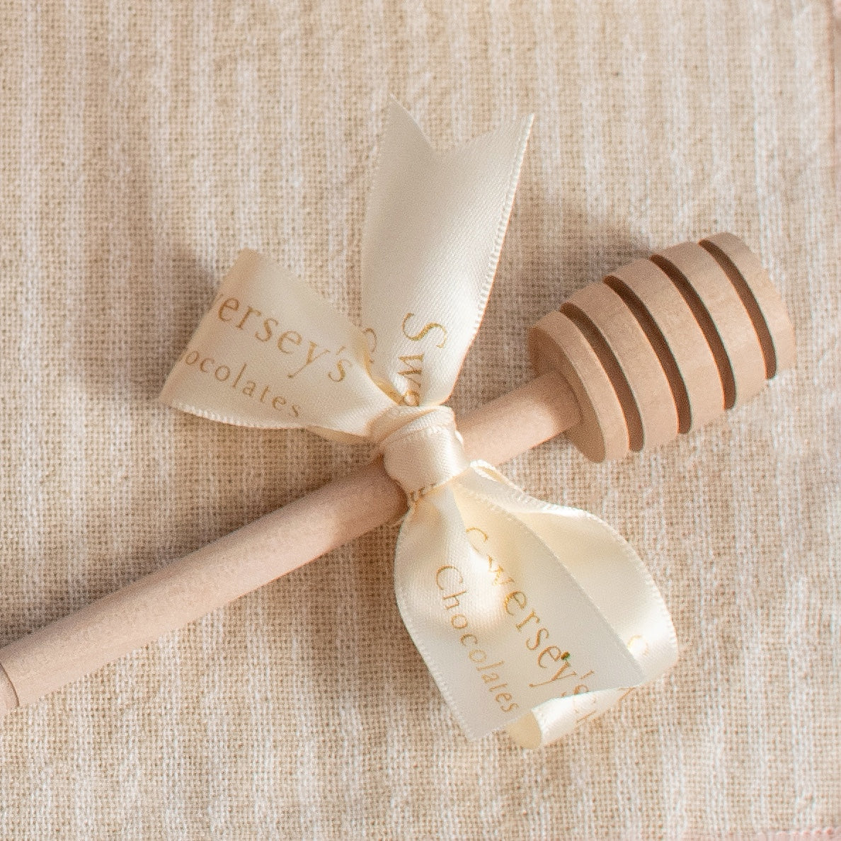 Kosherline wooden honey dipper tied with a cream-colored ribbon on a textured beige fabric background